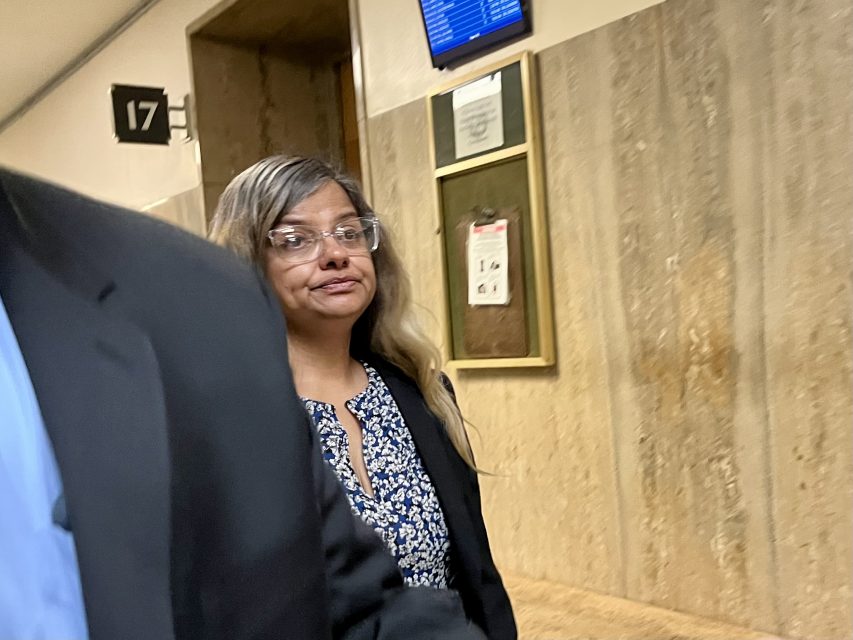 A woman wearing glasses and a patterned blouse walks down a hallway with a serious expression.