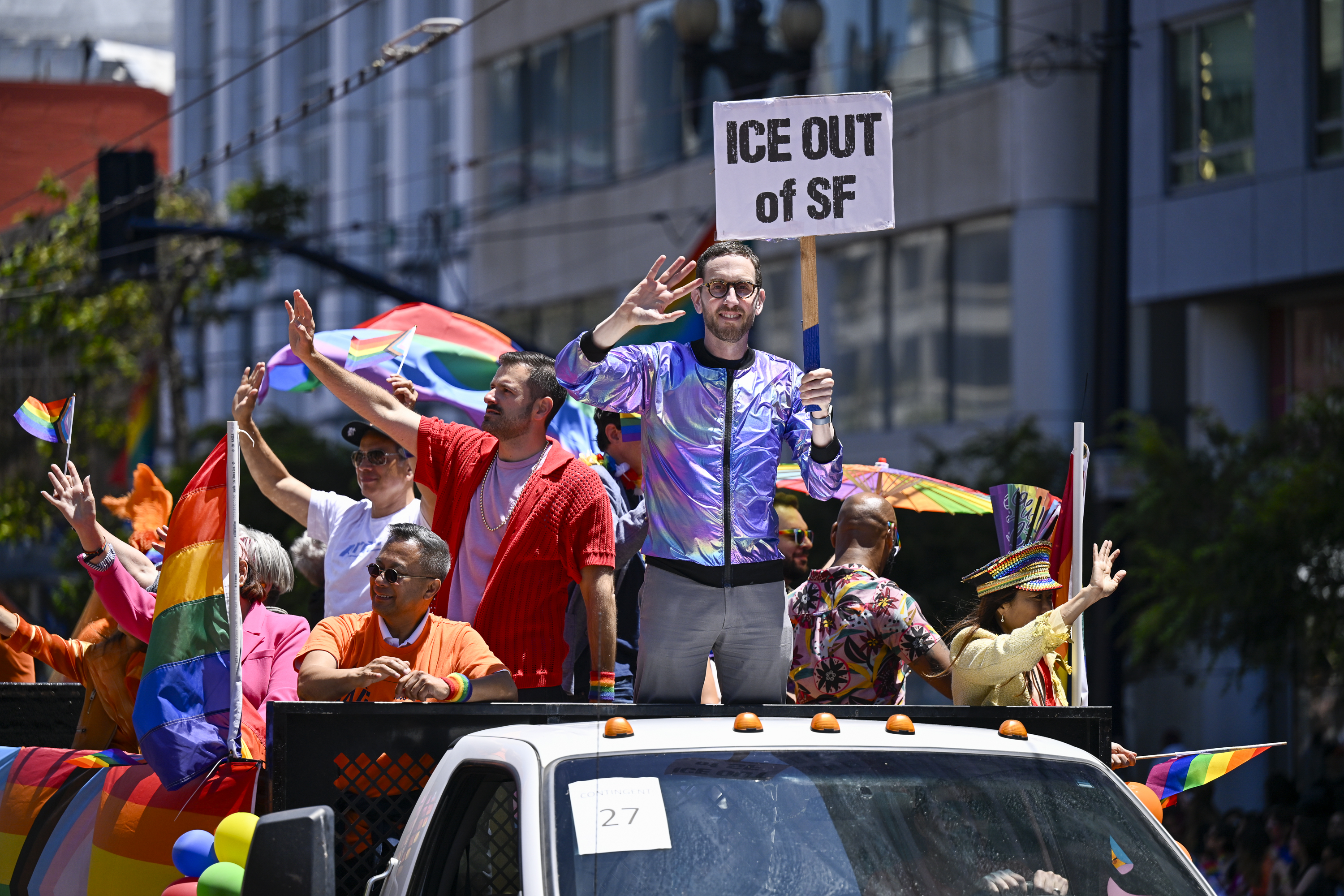 A bearded man in an iridescent jacket and glasses waves with one hand as he holds a sign in the other. The sign reads, "ICE OUT of SF." The man stands on the back of a flatbed truck decorated with rainbow flags alongside other people who are dressed in bright colors and  also waving to the unseen crowd.