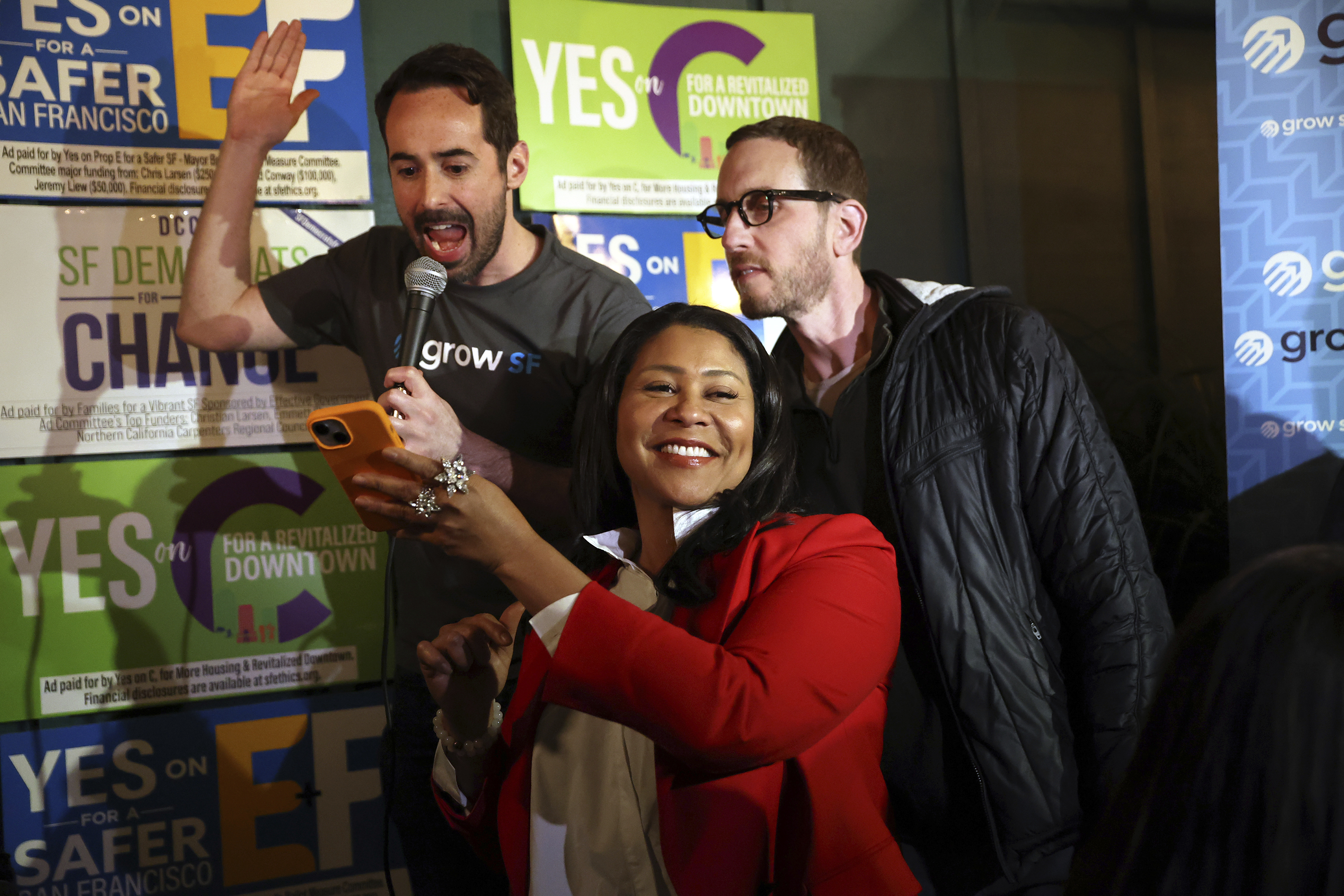 A middle-aged African American woman in a red blazer smiles as she holds up her phone for a man behind her. The man, holding a microphone, appears to be reading aloud to an unseen crowd. Another man, who has a beard and wears glasses, also leans into look at the phone. Behind them are campaign signs that read, "Yes on C for a Revitalized Downtown" and "SF Democrats for Change."