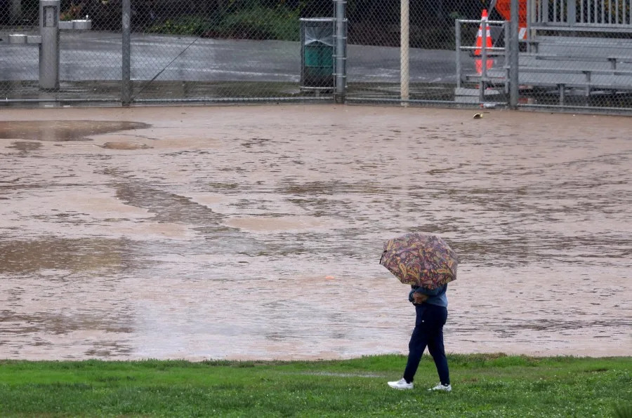 LOS ANGELES, CA – NOVEMBER 15, 2025 — A woman walks past a flooded baseball diamond in Downey Recreation Center Park in Los Angeles on November 15, 2025. (Genaro Molina/Los Angeles Times via Getty Images)