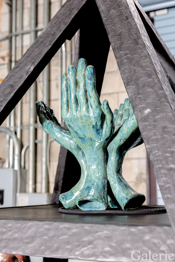 Sculpture of multiple hands reaching upward, crafted from greenish material, displayed in an industrial setting.