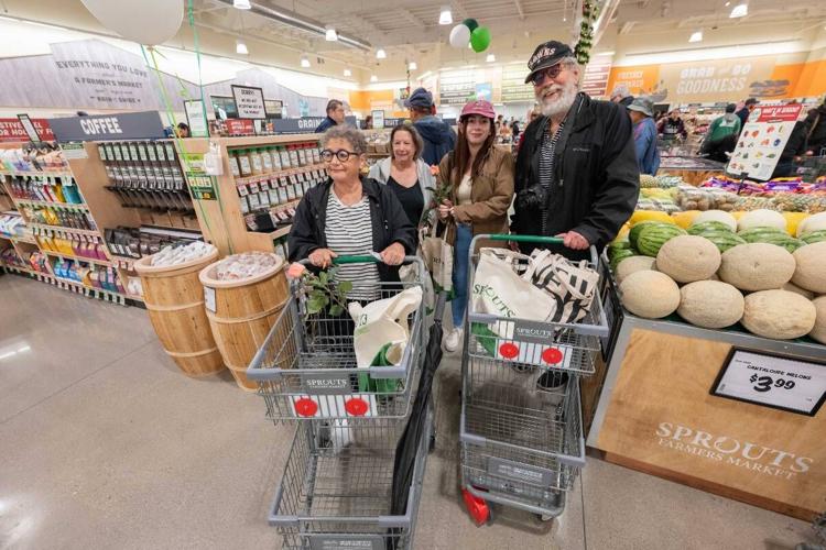 Four shoppers with carts stand near a display of cantaloupes inside a Sprouts store, holding tote bags and roses.