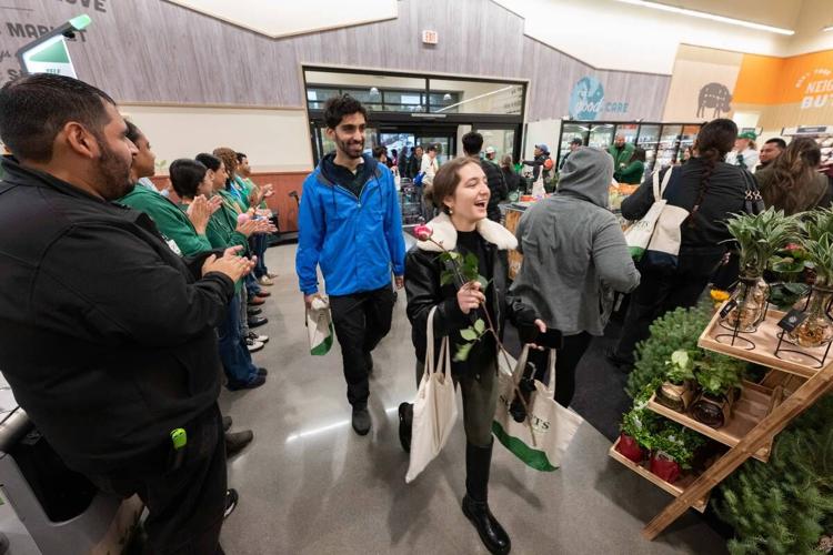 Two shoppers walk into a Sprouts store holding tote bags and roses while employees form a line and clap.