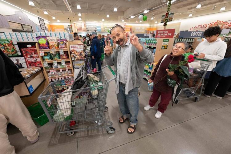 A man standing beside a shopping cart flashes two peace signs while a woman behind him holds a bouquet of roses inside a Sprouts store.