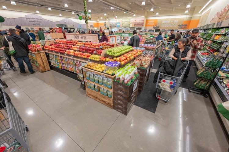 A wide view of a Sprouts produce section with shoppers pushing carts near displays of apples, citrus, and packaged snacks.