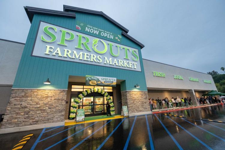 A wide view of a Sprouts Farmers Market storefront with “Now Open” signs and a balloon arch at the entrance, as a long line of people waits outside on the wet pavement.