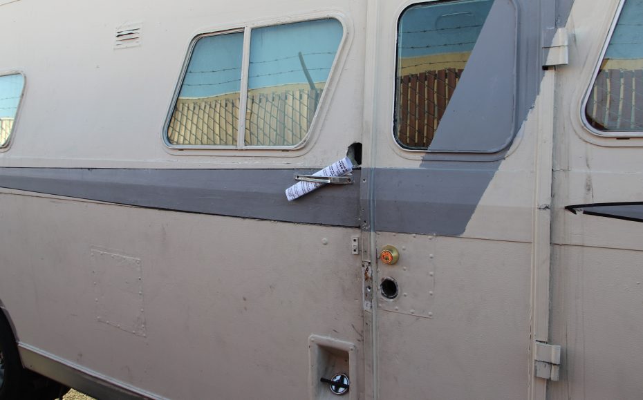 A rolled-up notice is wedged in the broken door lock of a beige RV, with a fence and barbed wire visible in the background.
