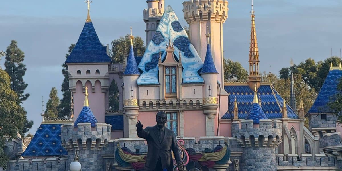 A bronze statue of Walt Disney holding Mickey Mouse stands in front of the Sleeping Beauty Castle with blue rooftops at Disneyland, surrounded by trees and partly cloudy skies.