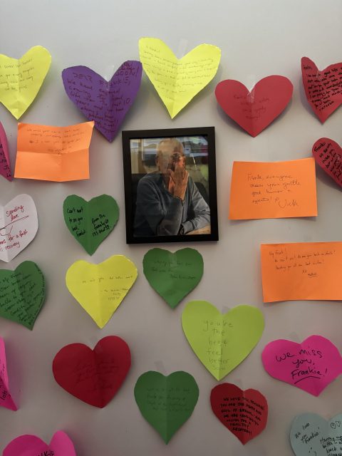 A framed photo of an elderly man is surrounded by colorful heart- and rectangle-shaped notes with handwritten messages on a wall.
