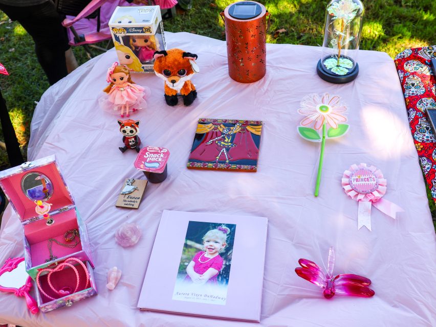 A table covered with a pink cloth displays toys, a photo album, a flower wand, a princess ribbon, a plush fox, a water bottle, and other colorful birthday gifts.