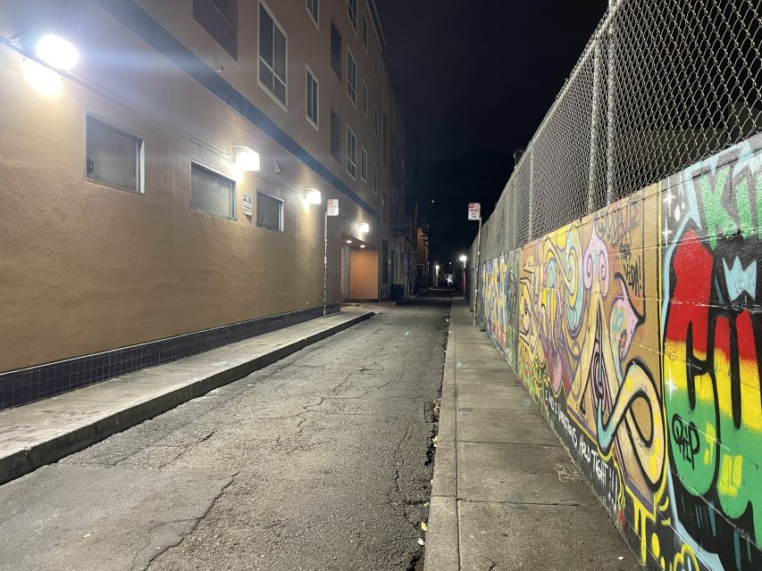 A narrow, empty alley at night is lit by wall lights, with a building on the left and a graffiti-covered fence on the right.