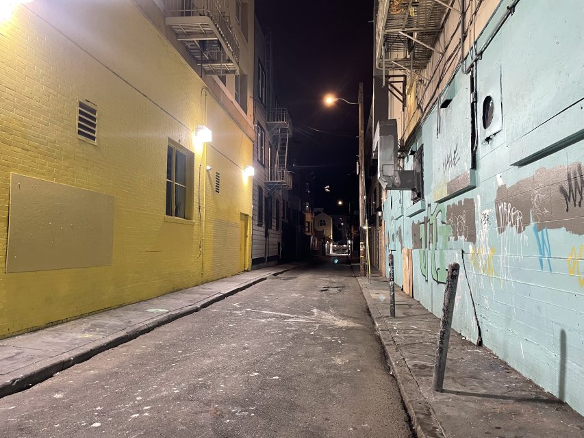 A narrow urban alley at night, lit by wall-mounted lights. One side is painted yellow, the other blue with graffiti. The alley is empty and lined with buildings.