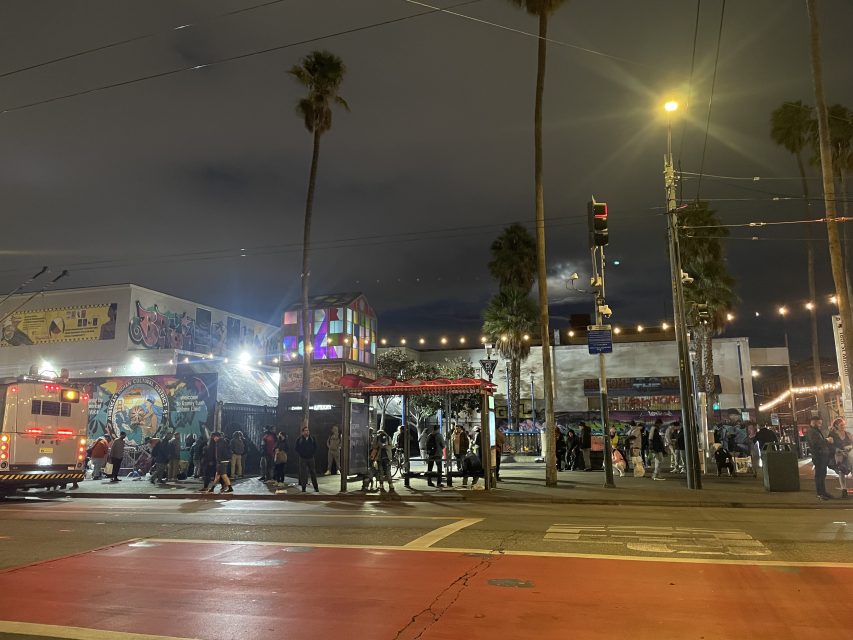 A busy street scene at night with people gathered outside colorful buildings and murals; palm trees, streetlights, and an emergency vehicle are visible.