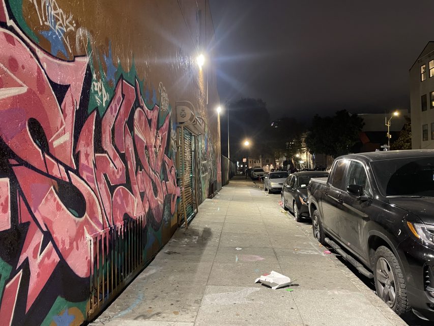 A city sidewalk at night with colorful graffiti on the left wall, parked cars on the right, and scattered litter on the ground under streetlights.