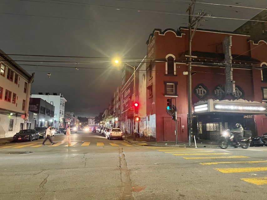 A nighttime urban street scene with cars, pedestrians, and a lit theater marquee on the corner under streetlights.