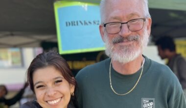 Two people wearing green sweatshirts stand side by side and smile at the camera at an outdoor market stall with a sign reading "DRINKS" in the background.