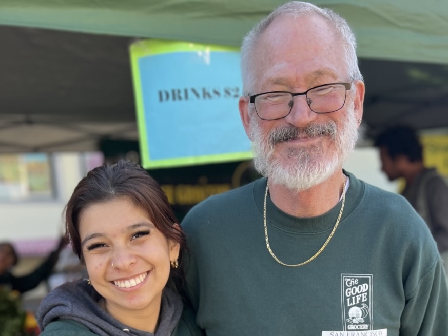 Two people wearing green sweatshirts stand side by side and smile at the camera at an outdoor market stall with a sign reading "DRINKS" in the background.