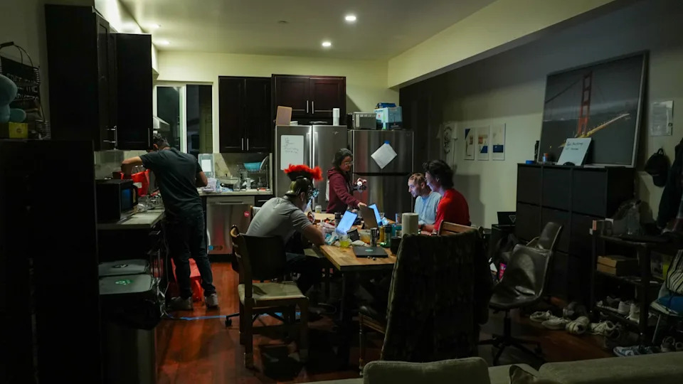 People work on laptops around a kitchen table at night, surrounded by snacks and drinks.