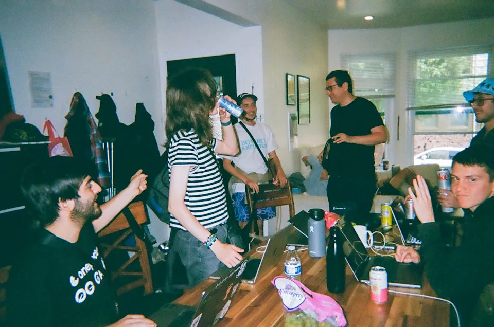 A group of people gather around a wooden table covered with laptops, drinks, and snacks in a casual home setting. Some talk and laugh while others work, creating a relaxed, social atmosphere.