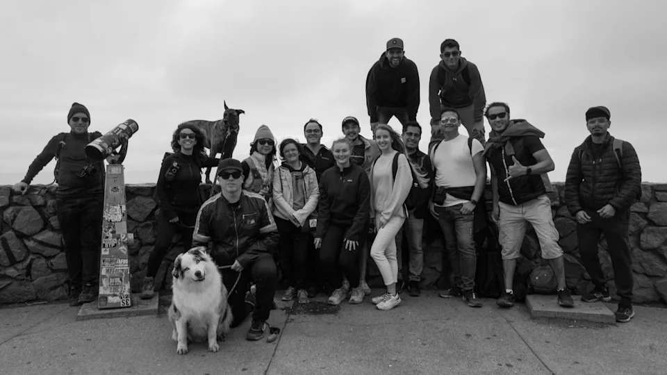 A group of hikers poses together on an overcast day at a scenic lookout point, smiling for a group photo beside a stone wall. A dog sits at the front, and another stands on the wall behind them.