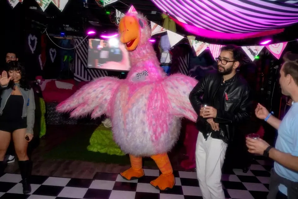 Partygoers dance under neon lights as a person in a giant pink-and-white bird costume entertains the crowd at a themed event, with attendees laughing and holding drinks on a checkered dance floor.