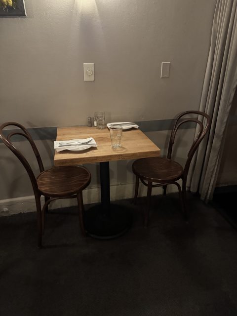 A small wooden table set for two with brown chairs, a glass of water, and utensils, positioned against a gray wall in a dimly lit room.