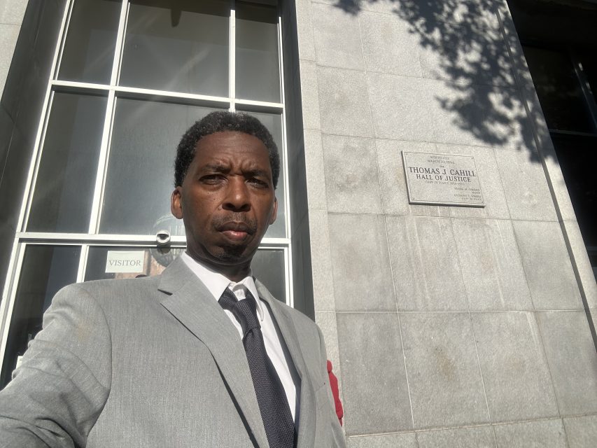 A man in a gray suit and tie stands outside a stone building near a window and a plaque reading "Thomas J. Cahill Hall of Justice.