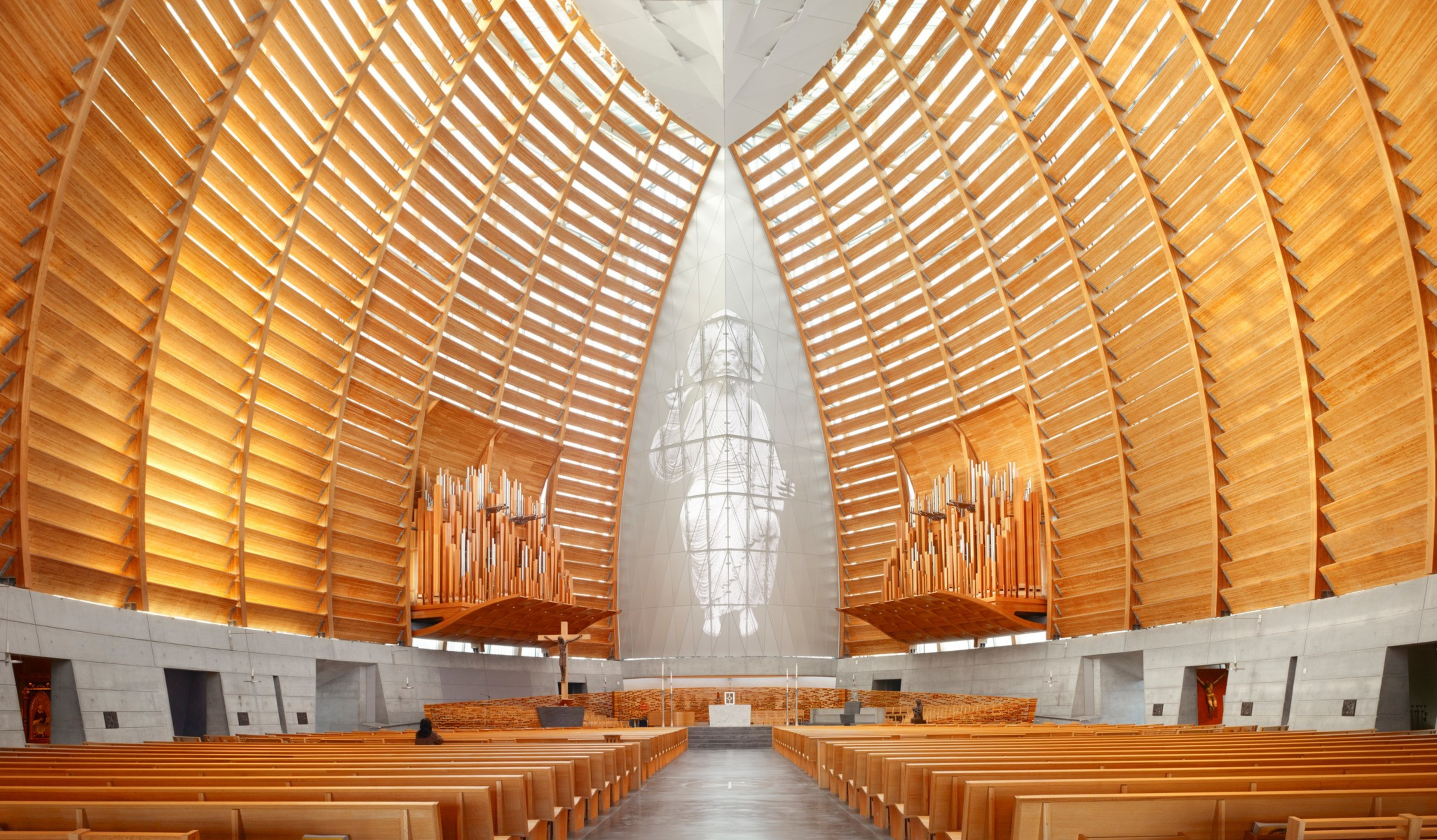 A spacious church interior features a wooden vaulted ceiling, two large organ pipes, wooden pews, a white altar, and a translucent statue at the center.