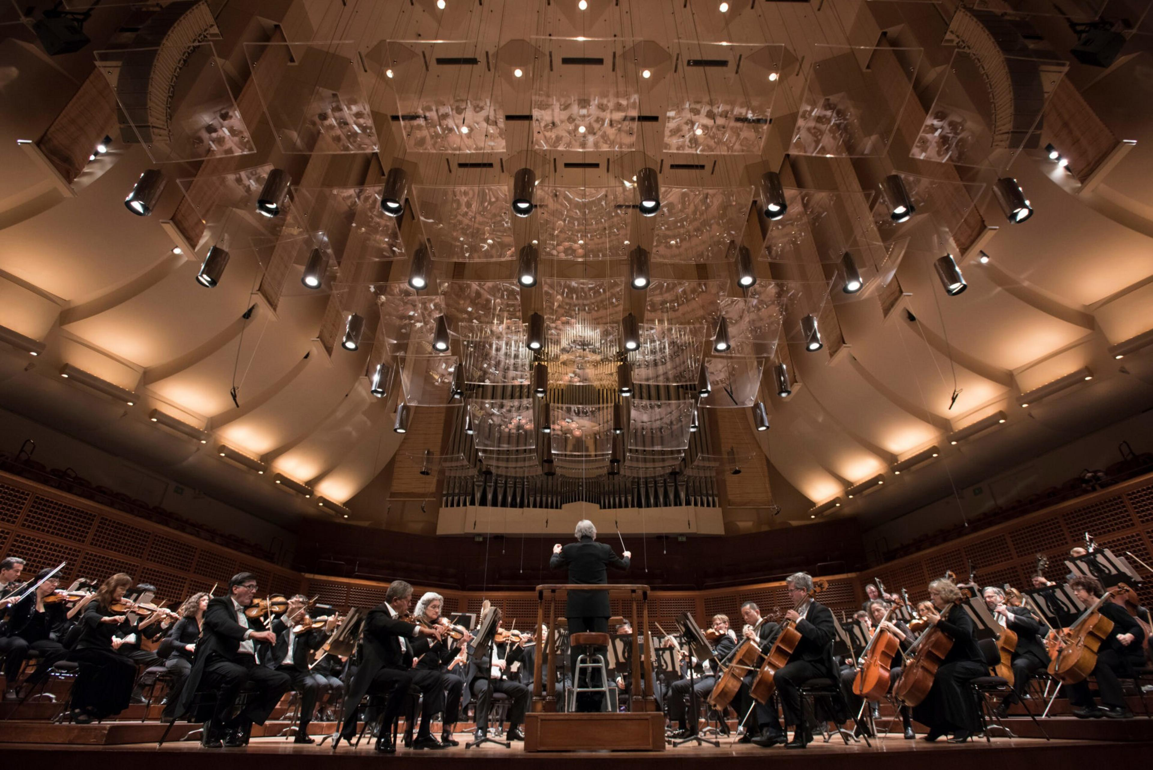 A symphony orchestra performs on stage in a grand concert hall with a conductor in front. The ceiling features numerous lights and acoustical panels.