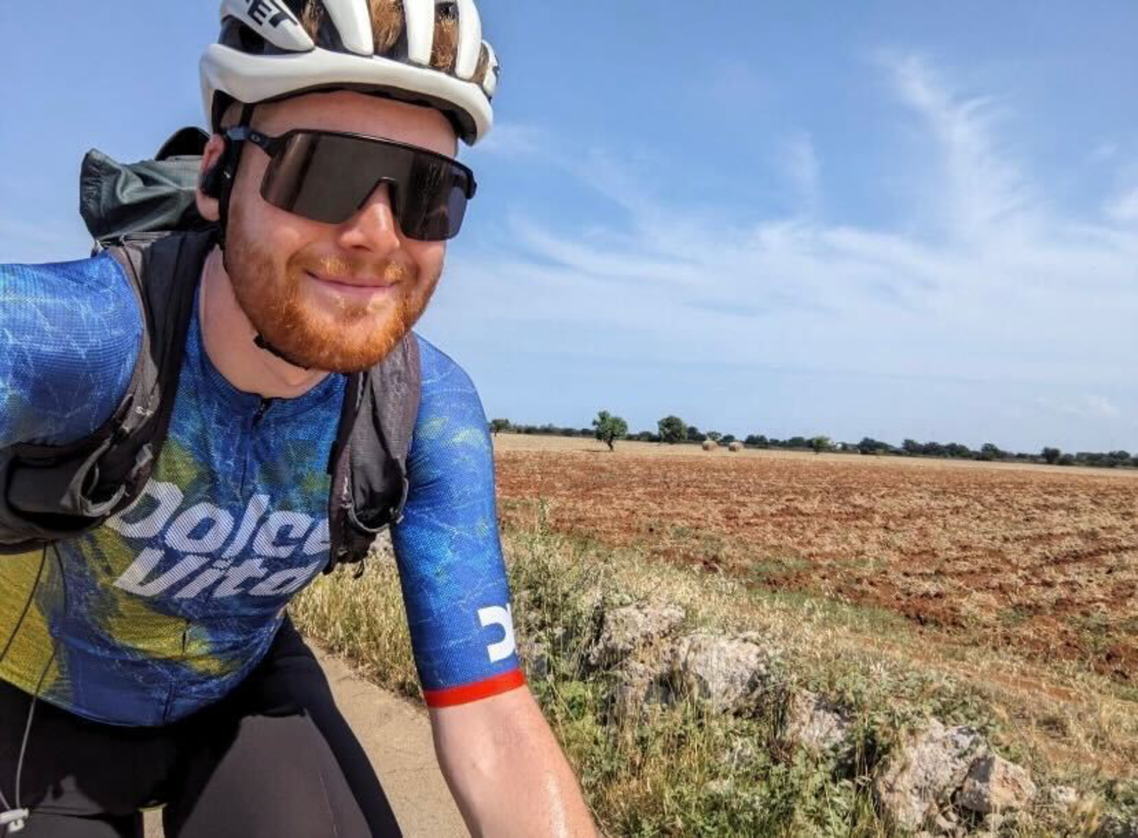 A cyclist wearing a white helmet, dark sunglasses, and a blue jersey smiles while riding near an open field under a clear, partly cloudy sky.