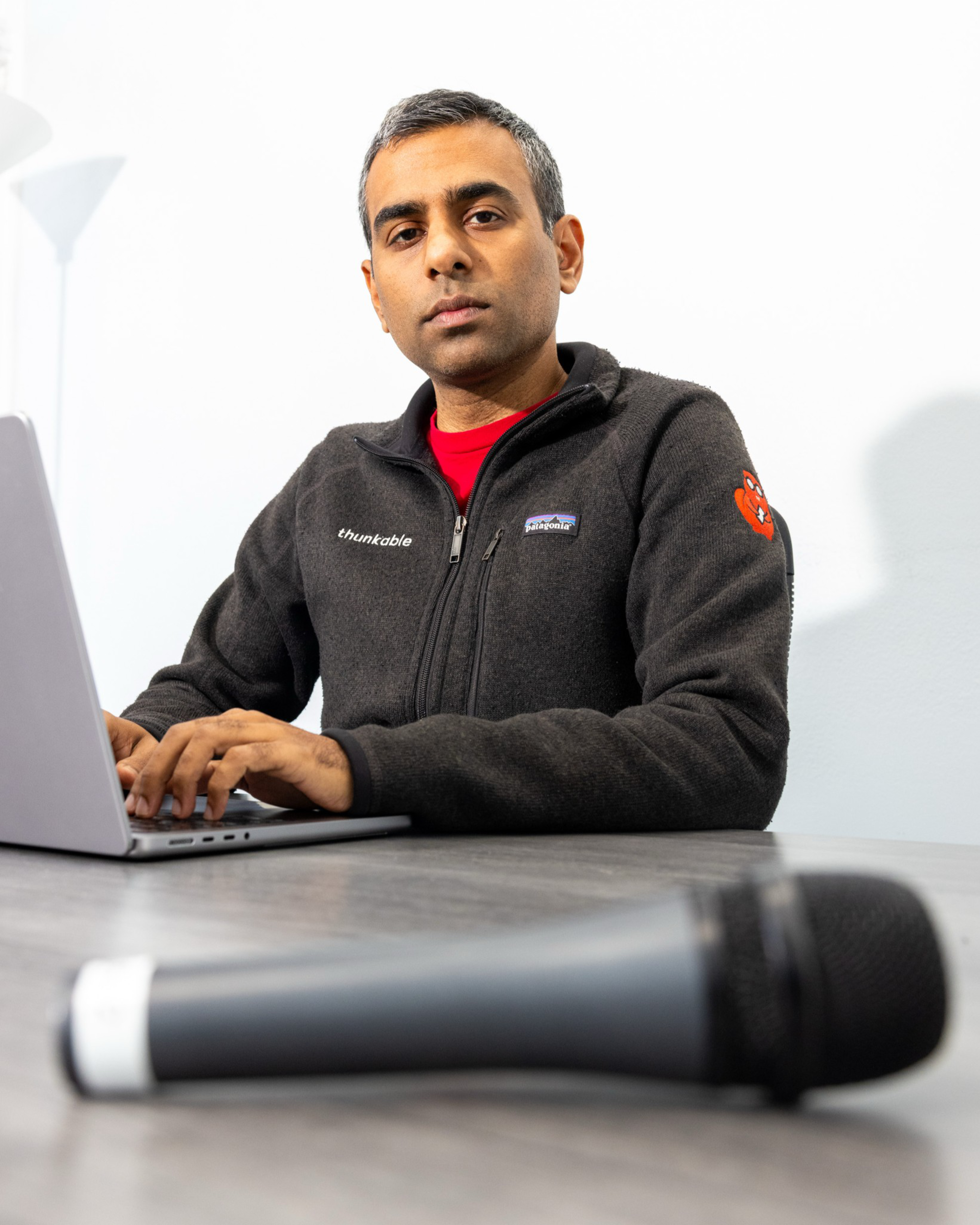 A man wearing a black fleece jacket with logos types on a laptop, sitting at a table with a microphone blurred in the foreground.