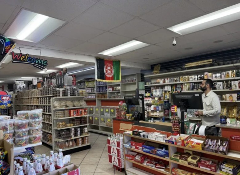 A man to the right stands behind a counter in a food mart lined with crowded shelves. An Afghan flag hangs from the ceiling.