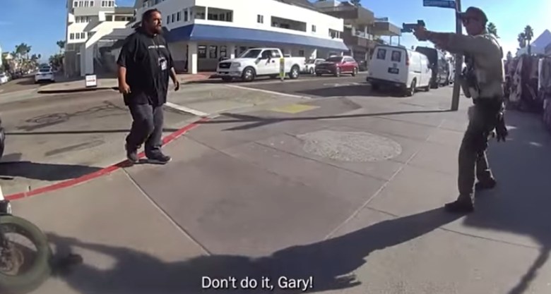 A large man approaches another man in a deputy's uniform who is holding a firearm. The are standing ona sidewalk near a city street.