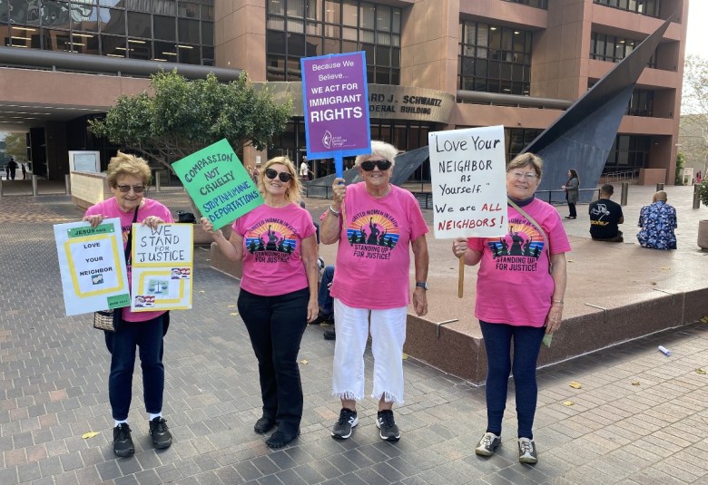 Four women dressed in pink T-shirts hold signs aloft showing their support for immigrants outside San Diego's courthouse.