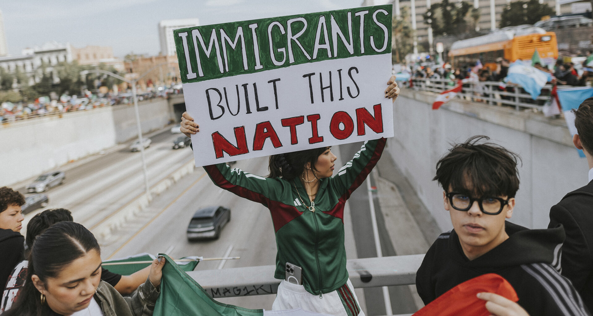 A person in a green tracksuit holding up a sign saying, “IMMIGRANTS UILT THIS NATION,” while next to a guard railing over a highway in downtown Los Angeles. In the background, cars can be seen driving on the road while crowds of protesters hold up signs and flags on both sides of the highway.