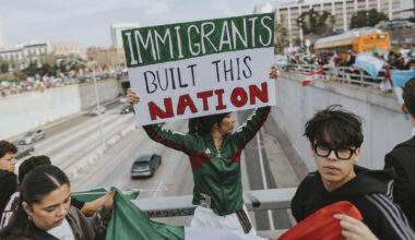A person in a green tracksuit holding up a sign saying, “IMMIGRANTS UILT THIS NATION,” while next to a guard railing over a highway in downtown Los Angeles. In the background, cars can be seen driving on the road while crowds of protesters hold up signs and flags on both sides of the highway.