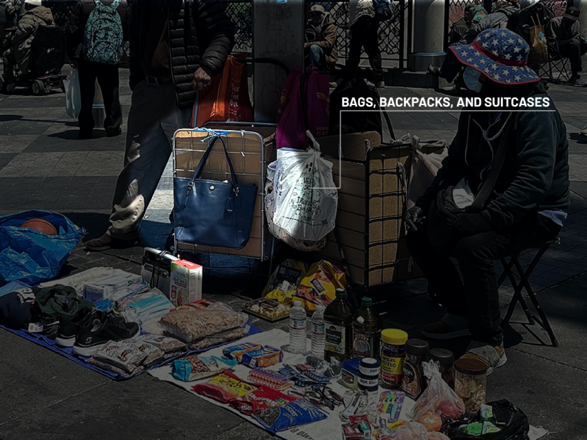 A street vendor sits beside bags, backpacks, and suitcases, with food items and goods displayed on a cloth on the pavement.