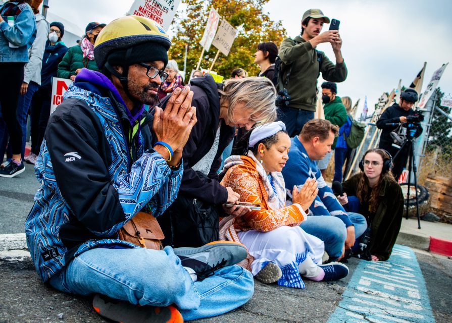 A diverse group of people kneel and sit on a street during an immigration protest, some with hands clasped as if in prayer, while others hold signs or use phones.