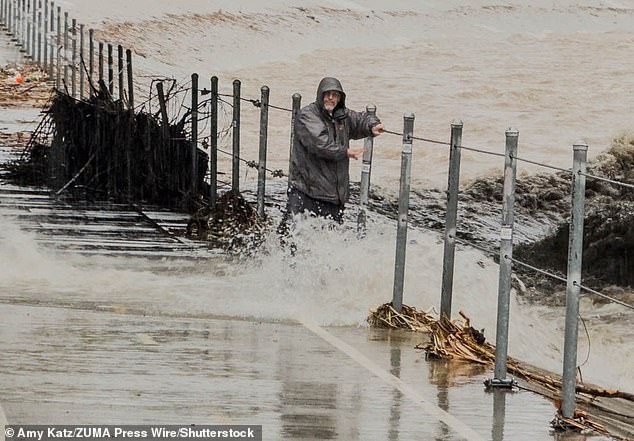 Thousands of California residents found themselves under evacuation orders on Christmas Eve, as disastrous floods and debris flows closed down roads. A man is pictured here hanging on to the railing for dear life after stepping into the path of a rushing culvert on Wednesday