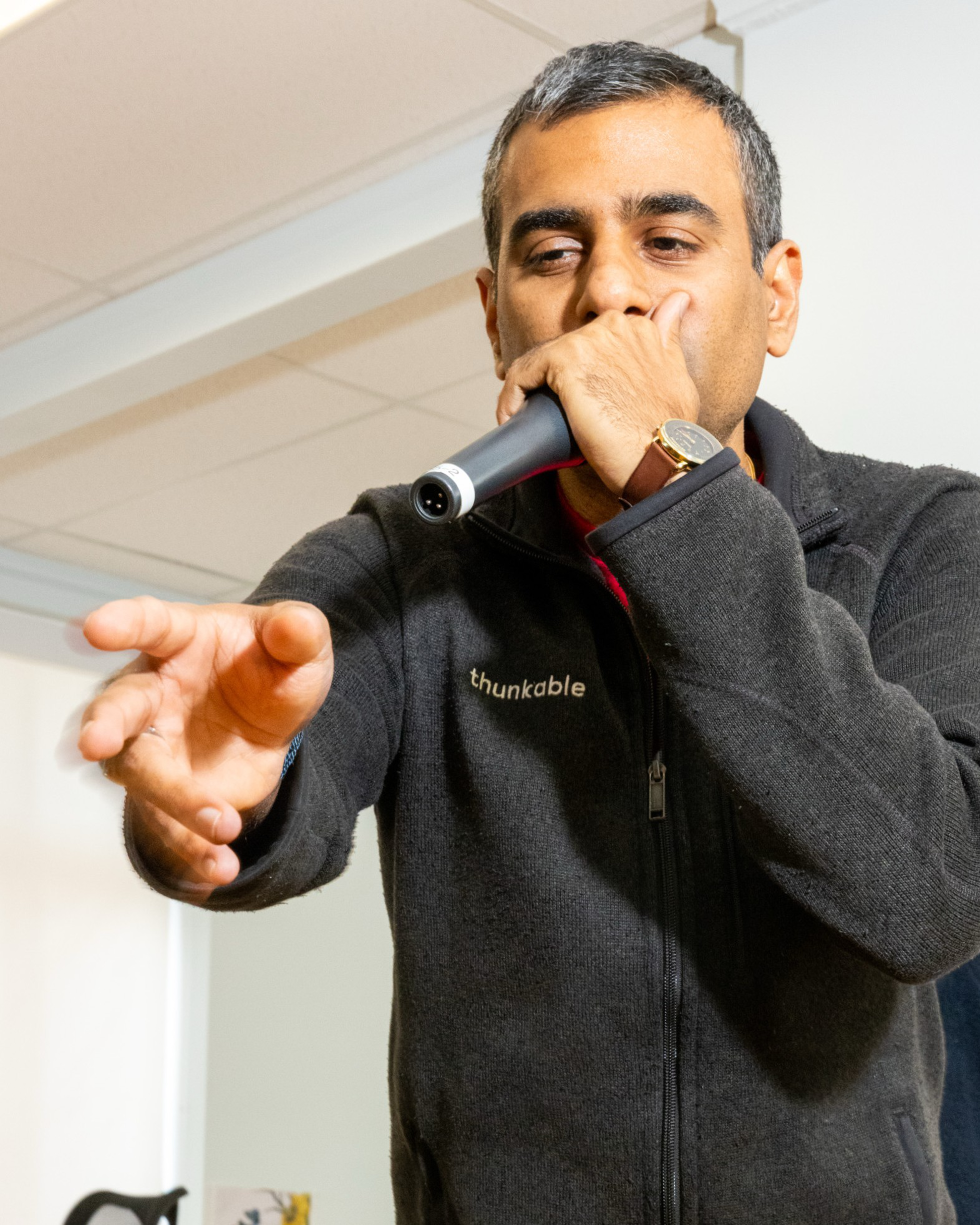 A man wearing a dark zip-up jacket with “thunkable” on it holds a microphone near his mouth and reaches forward with his other hand.