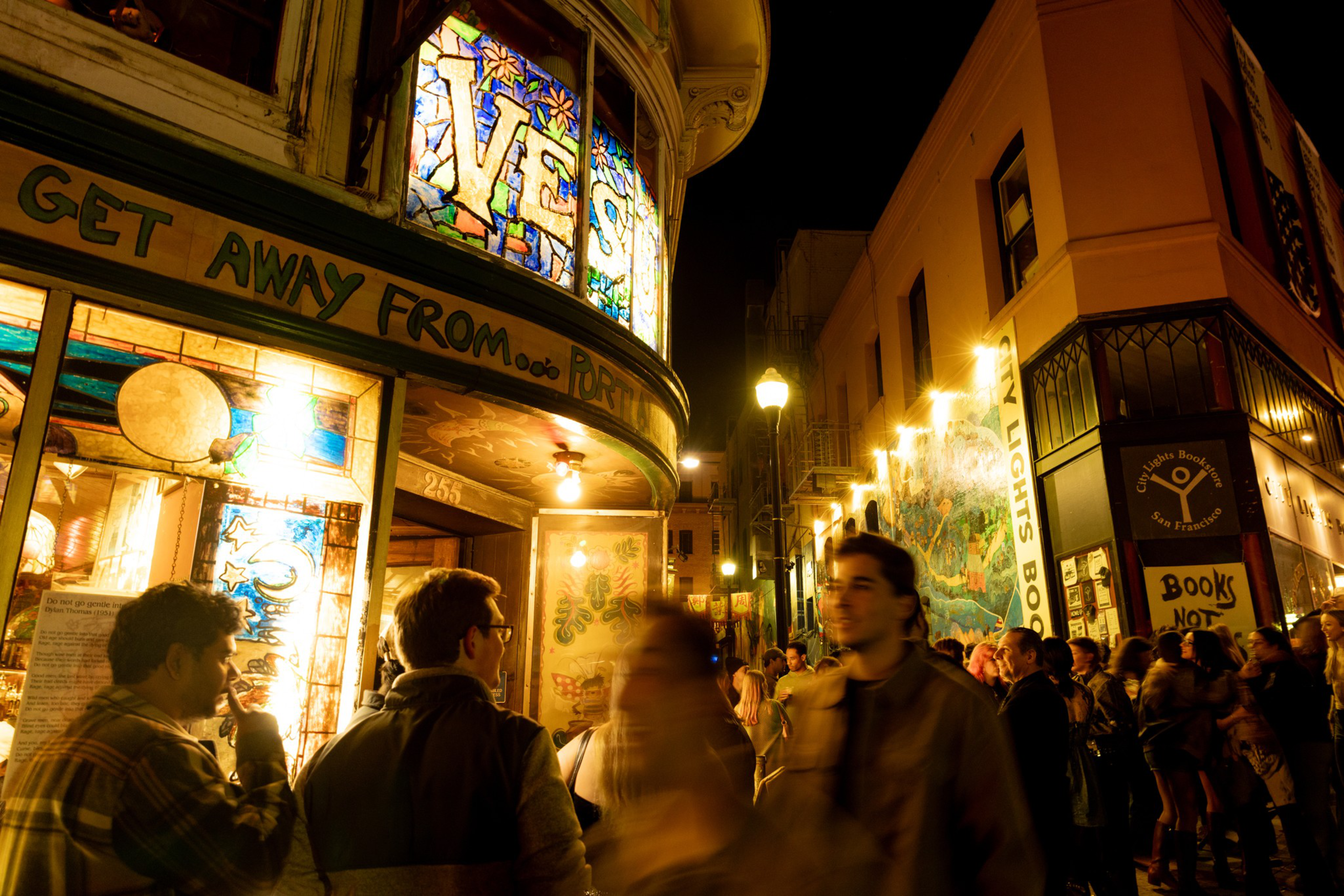 A lively night crowd gathers outside a brightly lit bookstore with stained glass windows and colorful signs in a narrow, warmly lit street.