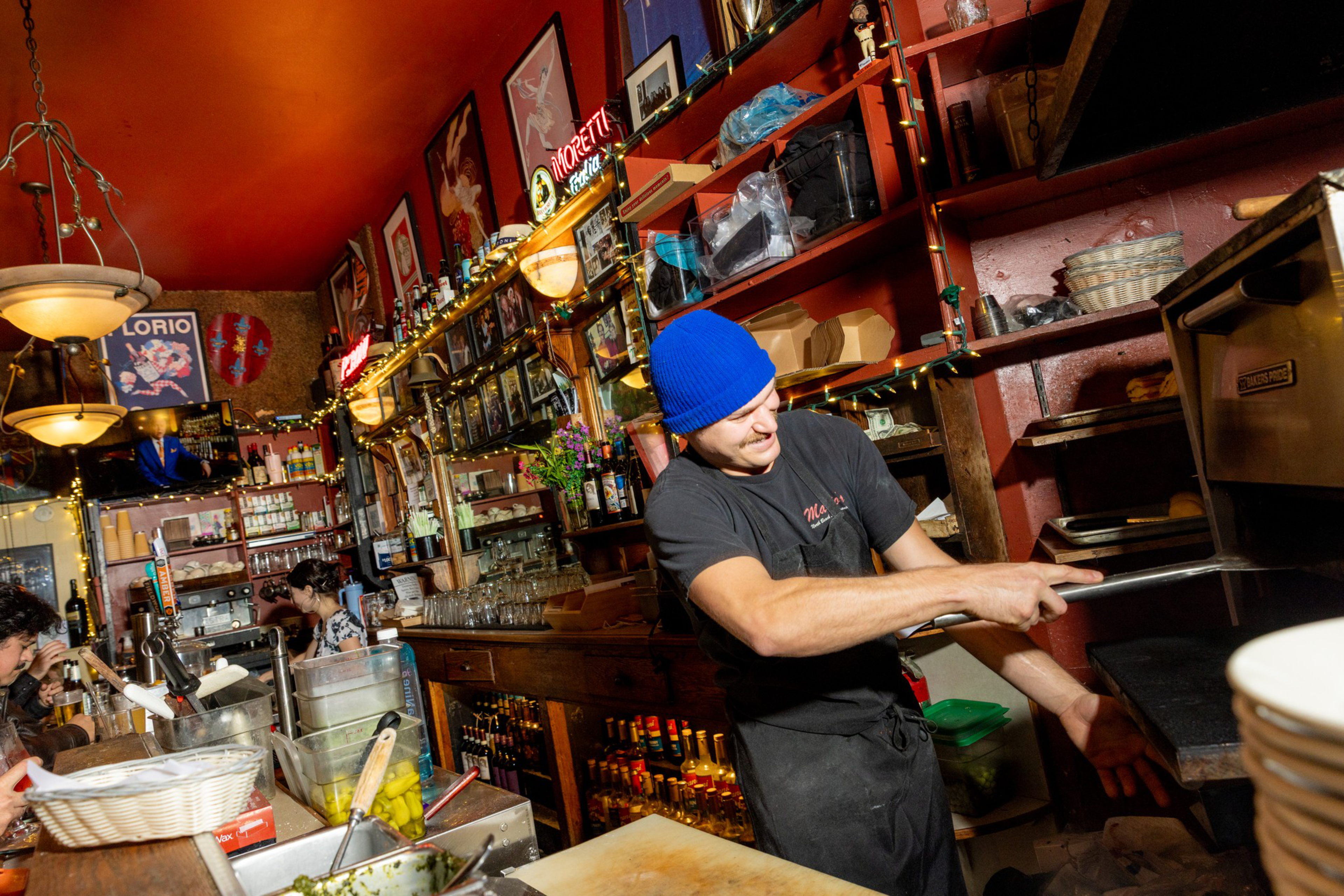 A man wearing a blue beanie and black shirt uses a peel to slide food into a pizza oven in a cozy, warmly lit restaurant with wooden shelves and decorations.