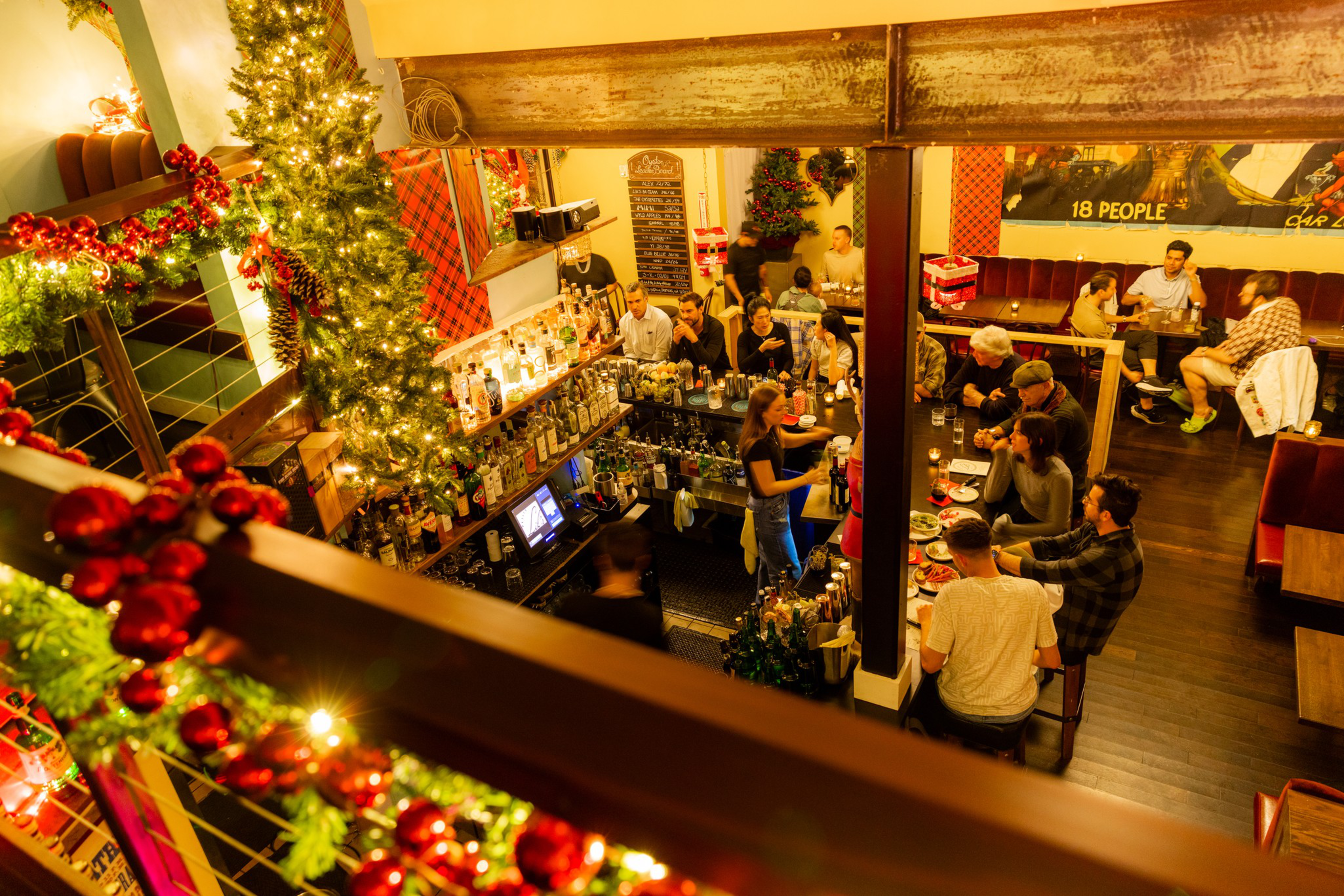 A warmly lit bar decorated with Christmas garlands and lights has patrons seated at the bar and tables, enjoying drinks and conversation.
