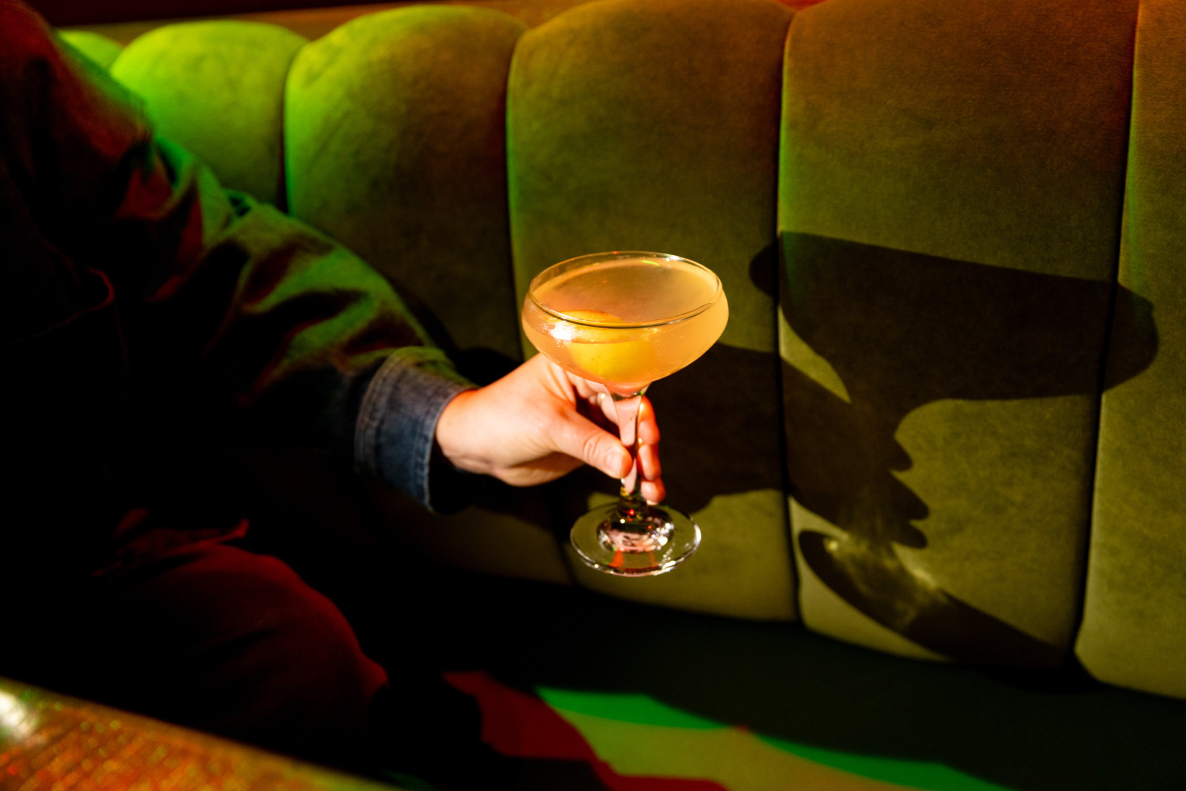 A hand holds a coupe glass with a pale yellow cocktail against a green upholstered bench, casting a clear shadow on the fabric.