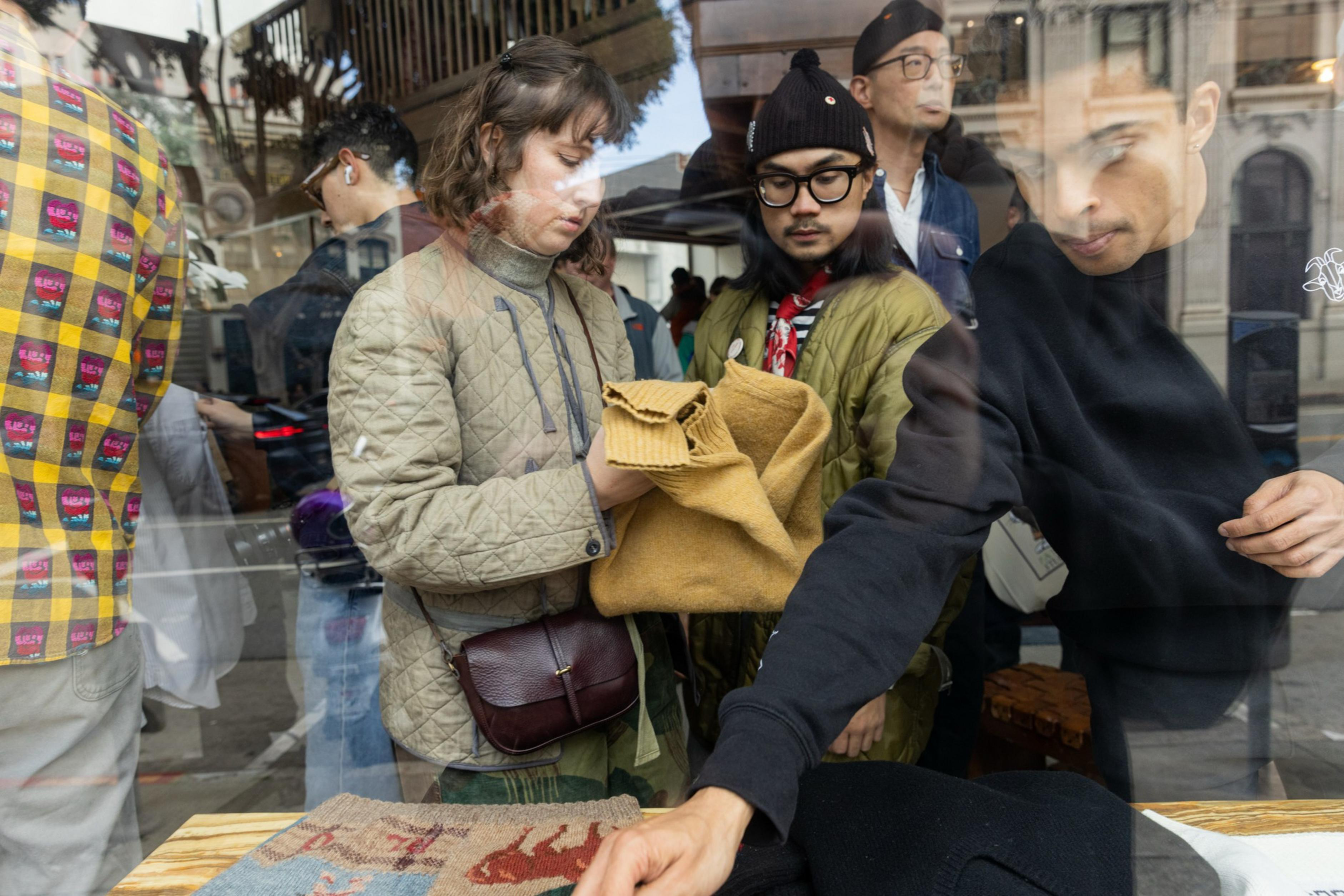 Three people inside a shop examine clothing, with one woman holding a folded mustard-colored sweater and another person reaching for a black garment.