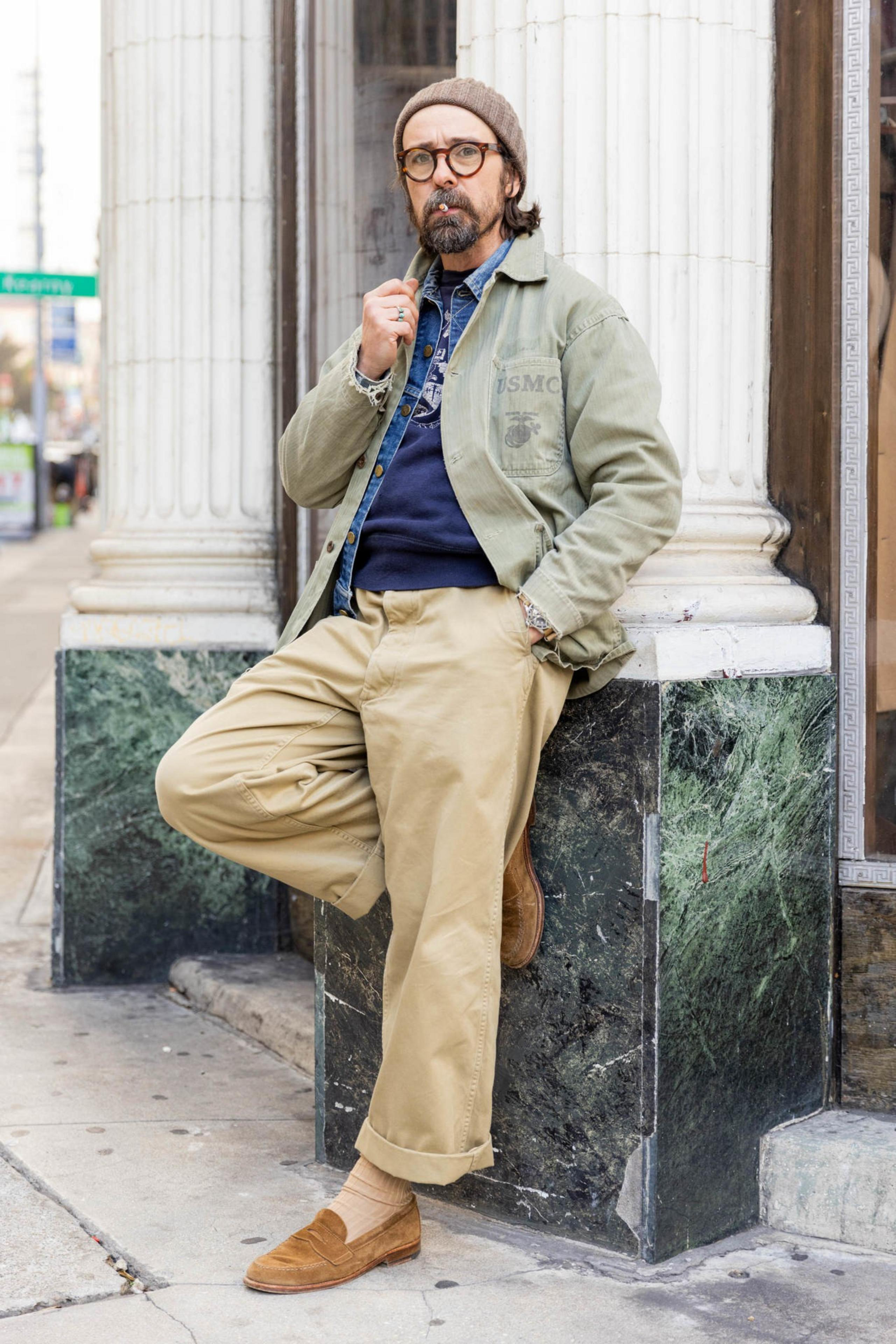 A man with glasses, a beard, and a beanie leans against a marble corner, wearing a green jacket, blue shirt, tan pants, and brown shoes.