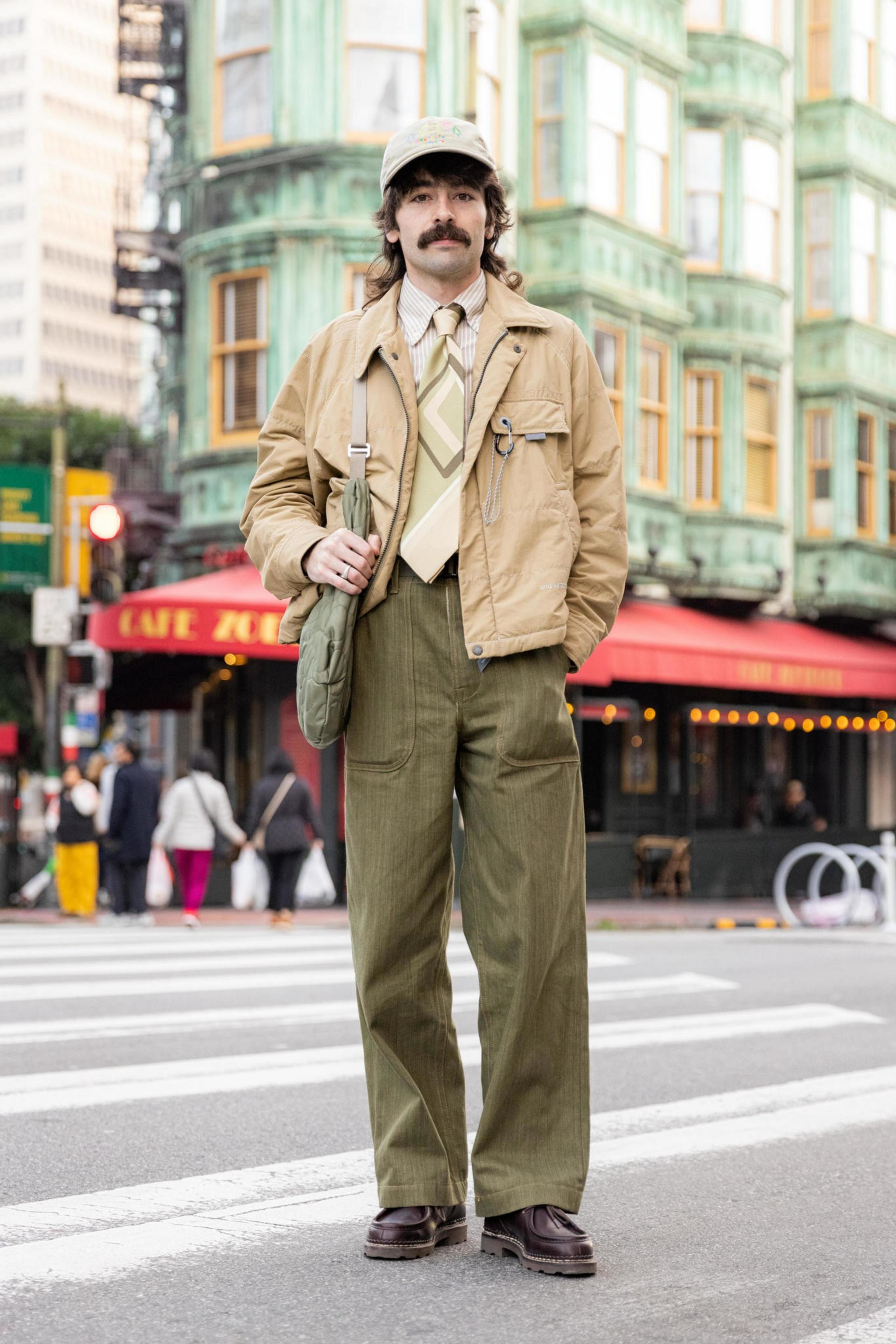 A man with a mustache wears a beige jacket, patterned tie, olive green pants, and a cap, standing on a city street crosswalk.