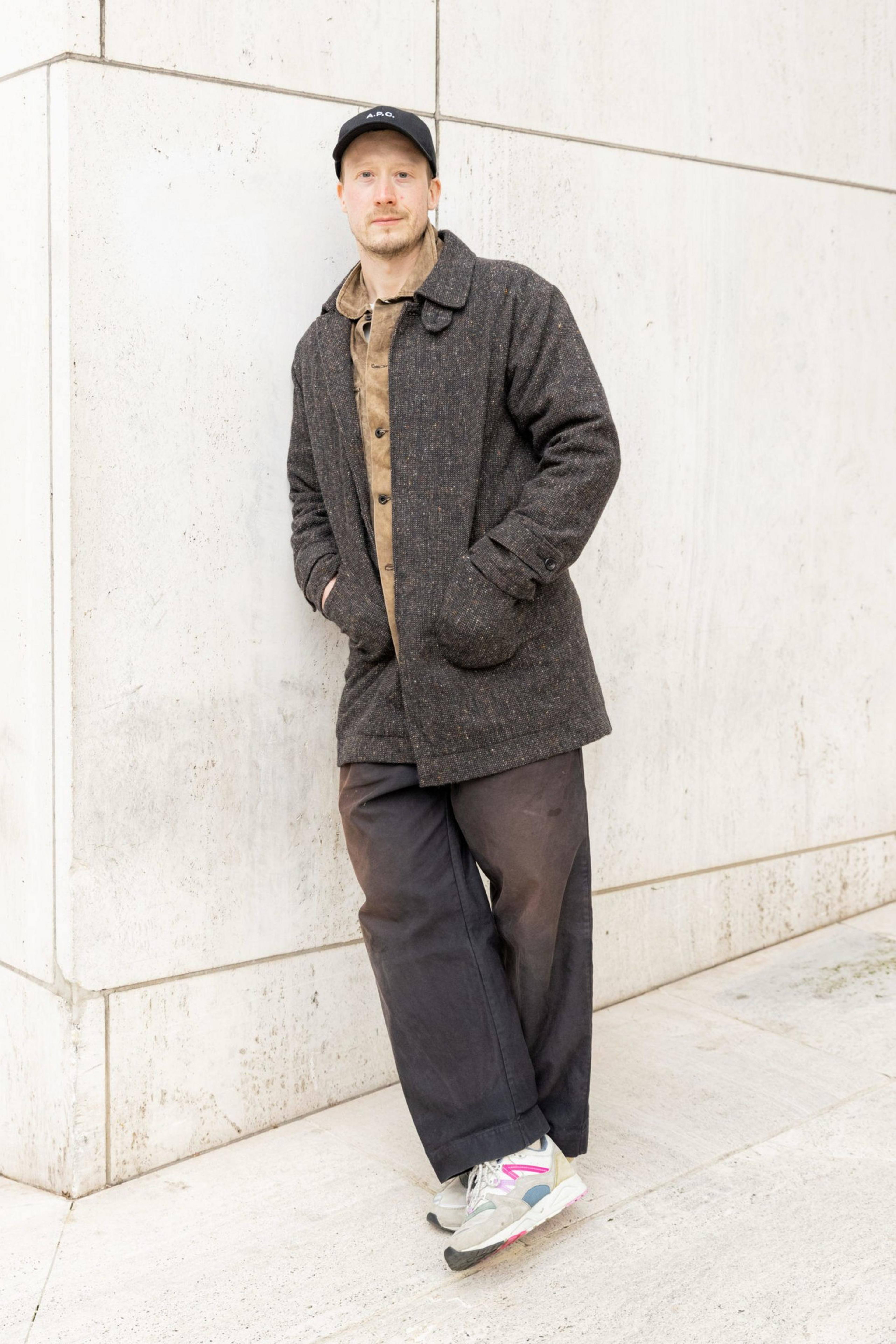 A man leans against a light stone wall wearing a dark gray coat, brown shirt, loose black pants, white sneakers with pink and blue accents, and a black cap.