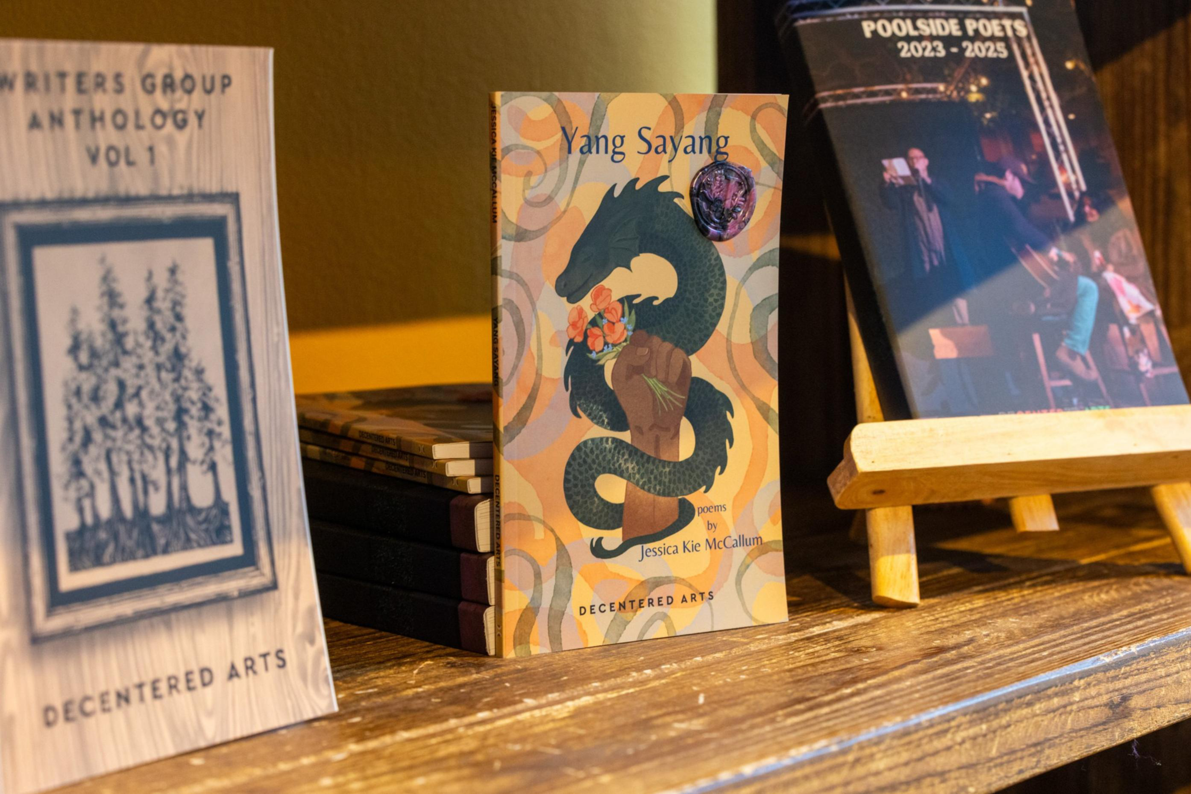Three books are displayed on a wooden shelf, with titles including “Writers Group Anthology Vol 1,” “Yang Sayang” by Jessica Kie McCallum, and “Poolside Poets 2023-2025.”