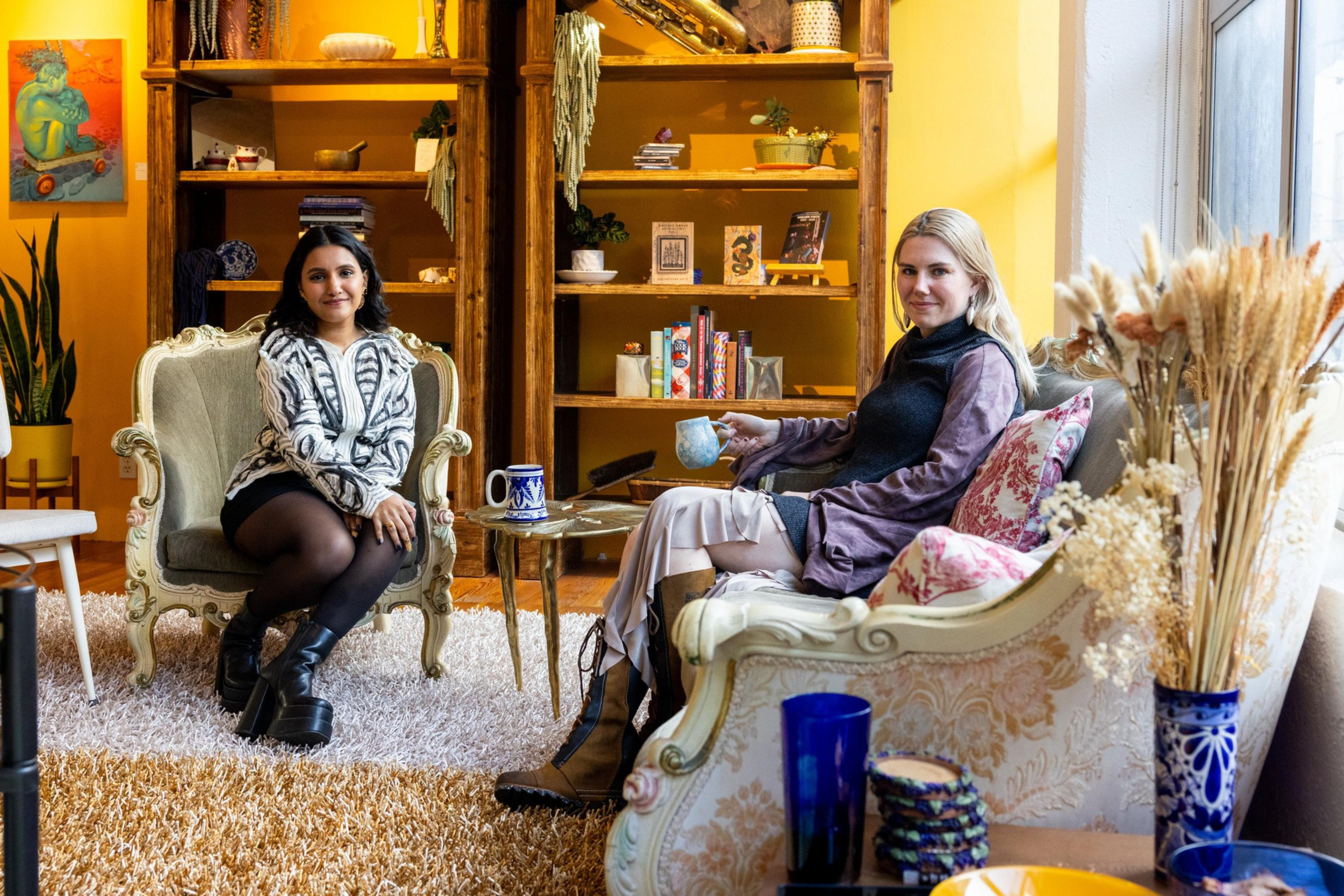 Two women sit in vintage armchairs in a cozy, warmly lit room with wooden shelves, plants, and decorative pottery around them.